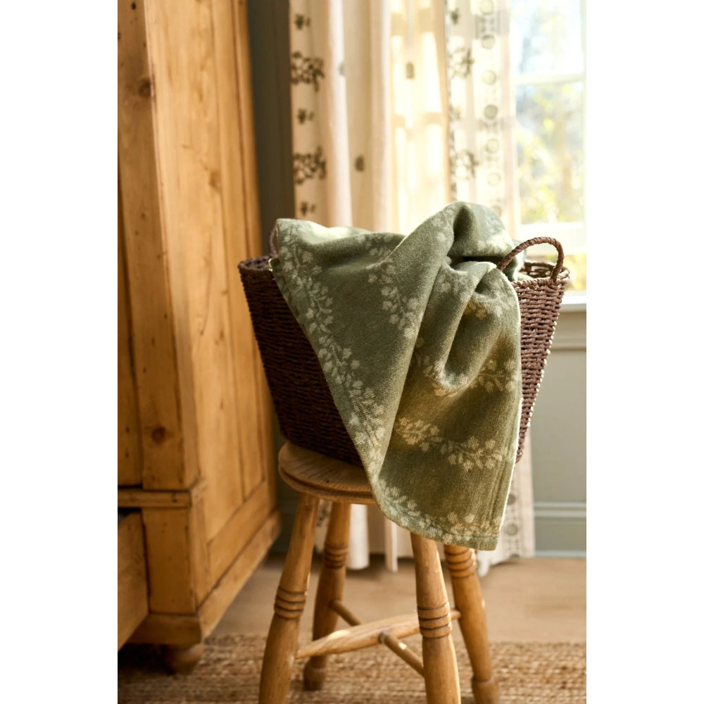 A sage green throw blanket with a cream floral pattern draped over a brown wicker basket on a wooden stool in a warm, rustic room.