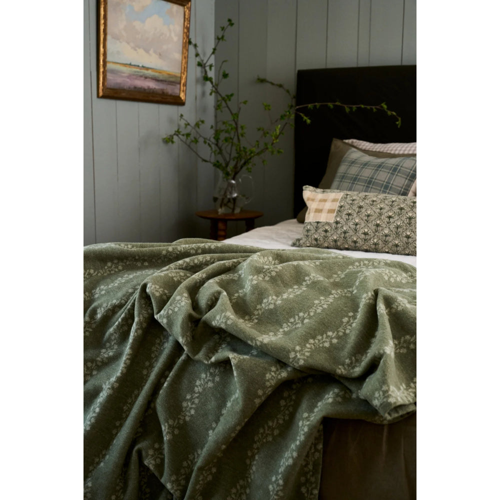 An olive green textured throw blanket with a white floral pattern draped over a bed in a styled bedroom setting.