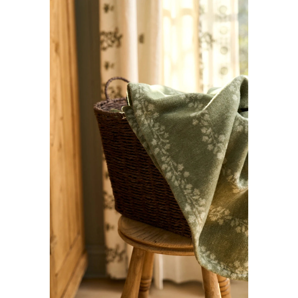 A sage green throw blanket with a white floral pattern draped over a dark brown woven wicker basket on a wooden stool in a softly lit room.