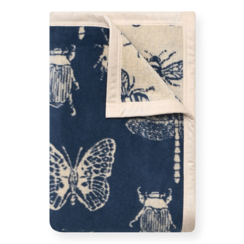 A folded blanket featuring a navy blue and cream indigo-style print of butterflies, moths, and beetles, with a cream border and a folded corner showing the reverse side.