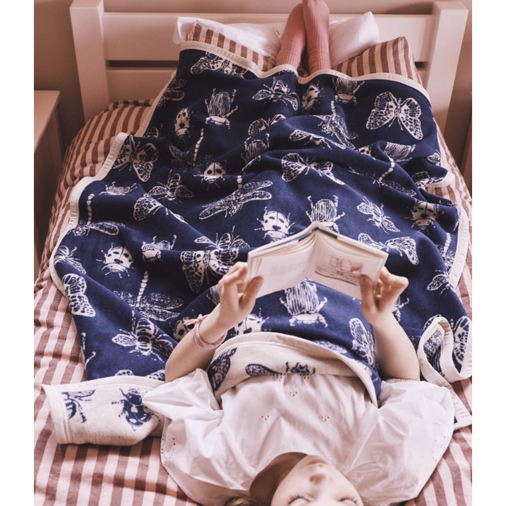 A person lying on a bed under a navy blue blanket printed with white butterflies and dragonflies, holding an open book. The bed has pink-and-white striped bedding, and the scene is photographed from above.
