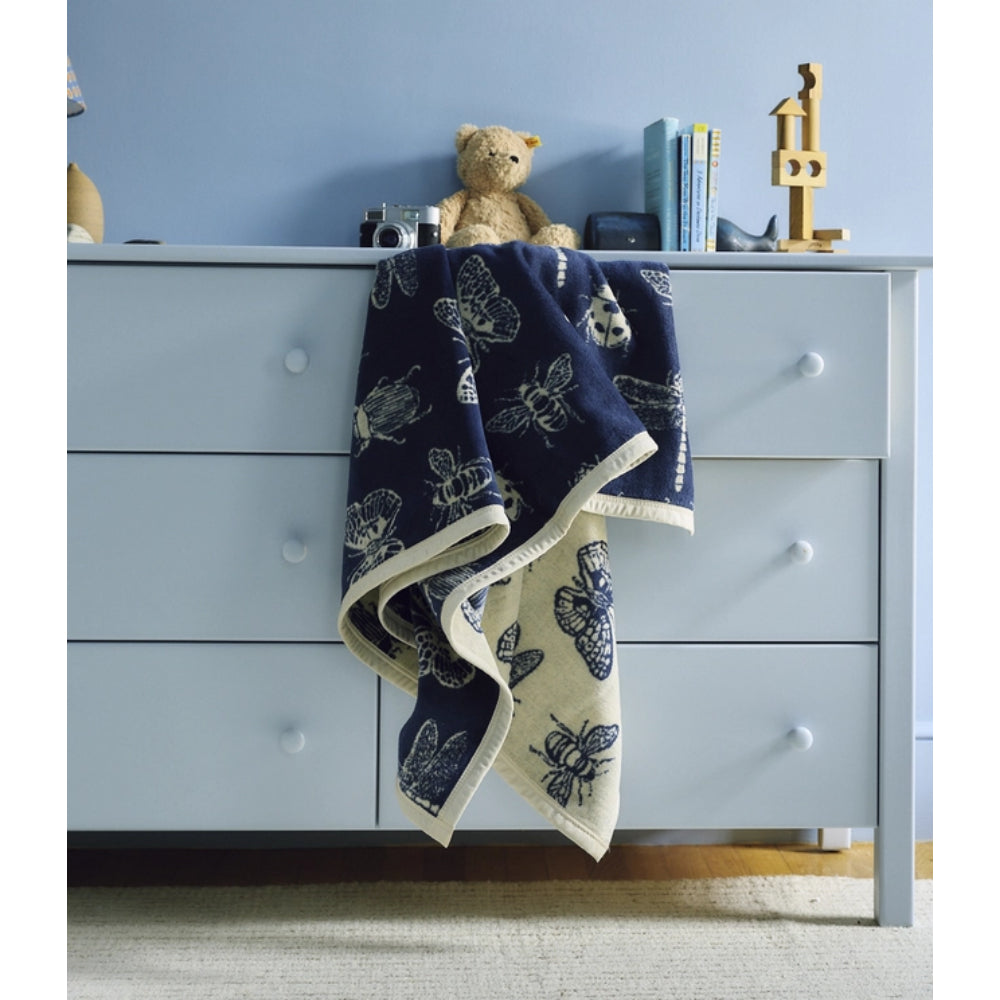 A navy blue baby blanket with a cream butterfly and bee print is draped over a light blue dresser in a nursery setting, with a teddy bear and books on top.