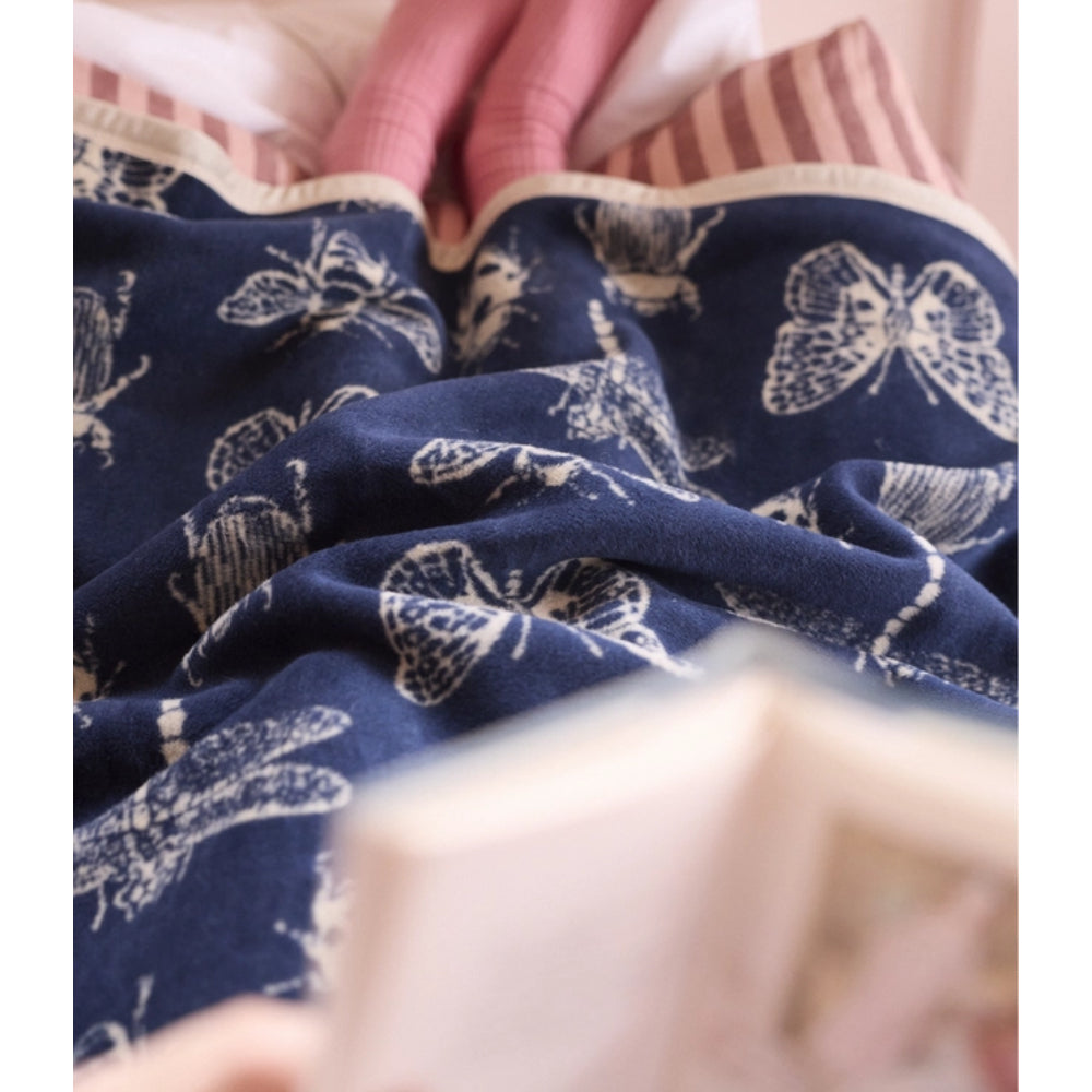 A close-up of a navy blue fabric item, likely a blanket or throw, printed with white butterfly and dragonfly motifs. The fabric is draped in soft folds, with a blurred pink striped textile and a light-colored object in the foreground.