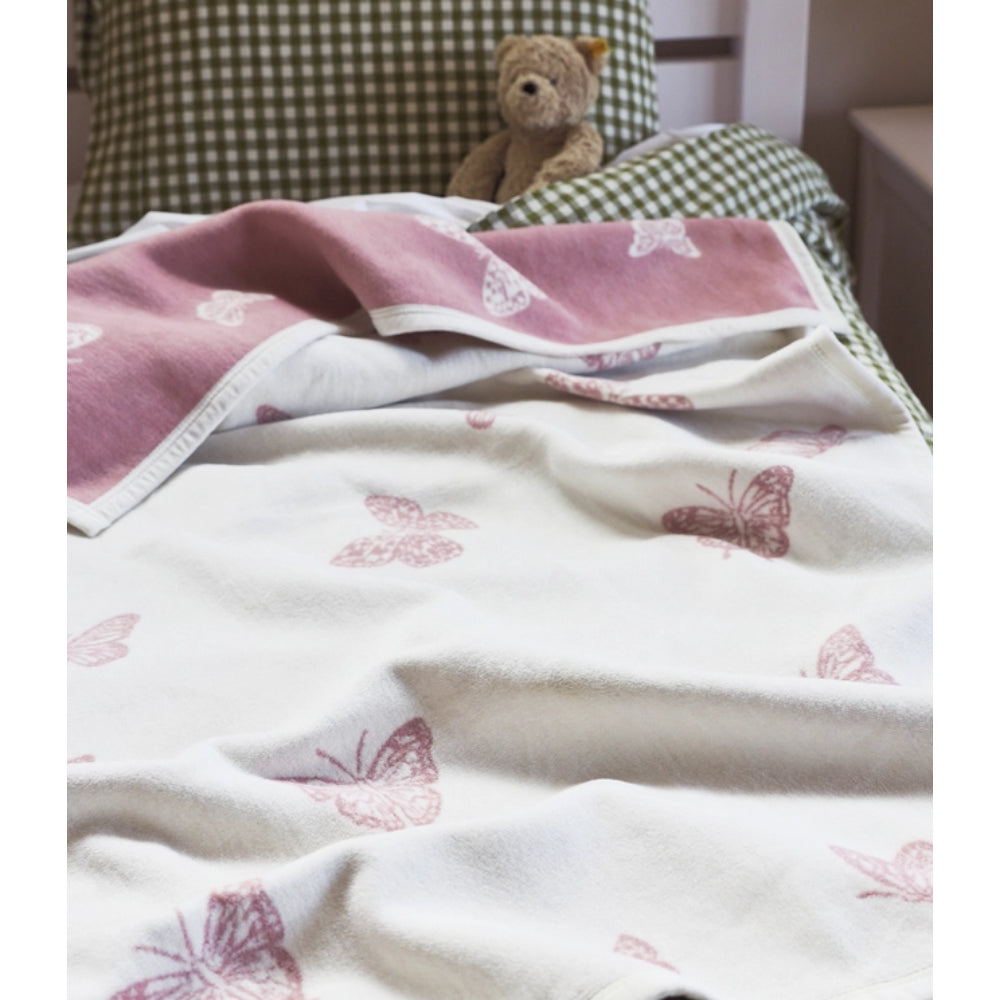 A soft white blanket with dusty pink butterfly prints draped over a bed, with a green-and-white checkered pillow and a small teddy bear visible in the background.