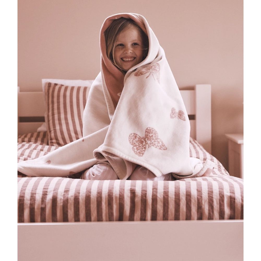 A smiling young girl sits on a bed wrapped in a soft white blanket with dusty pink butterfly prints. The room has a warm, muted pink color palette with matching striped bedding and pillows.