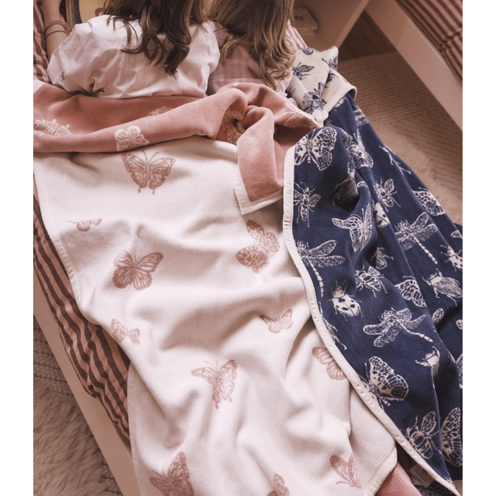 Two children lying under a soft blanket in a cozy bedroom setting. The blanket features a dual-sided design: one side is off-white with dusty pink butterfly prints, and the other side is navy blue with white dragonfly prints.