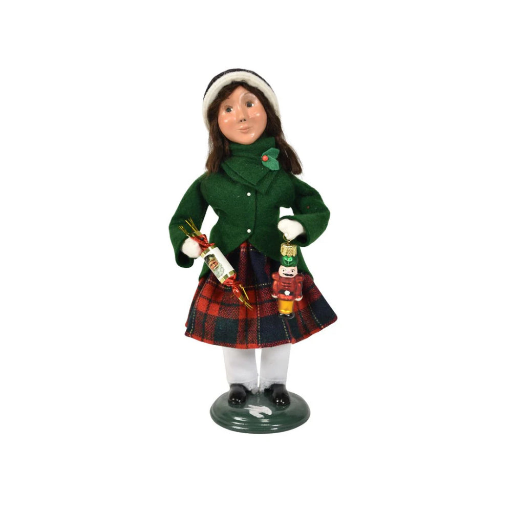 A figurine of a caroling girl wearing a green coat and red plaid skirt on holding a nutcracker ornament and holiday popper on a white background