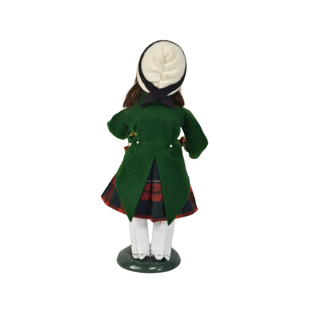 A back photo figurine of a caroling girl wearing a green coat and red plaid skirt on holding a nutcracker ornament and holiday popper on a white 
