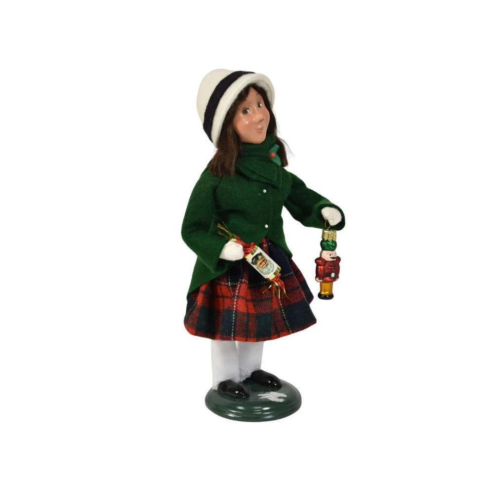 A right side angled photo of a figurine of a caroling girl wearing a green coat and red plaid skirt on holding a nutcracker ornament and holiday popper on a white background