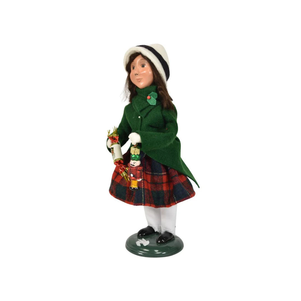 A left side angled photo of a figurine of a caroling girl wearing a green coat and red plaid skirt on holding a nutcracker ornament and holiday popper on a white background