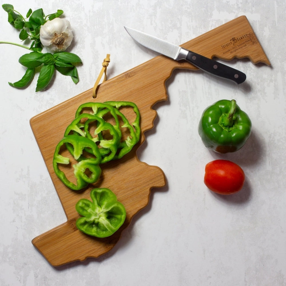 Wooden cutting board with sliced green peppers, a whole green pepper, and a red tomato on a light gray surface.