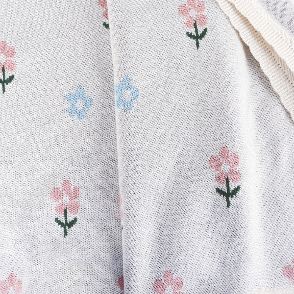 Close-up of a white knit fabric with a delicate pattern of small pink and light blue flowers with green stems, featuring a ribbed cream-colored trim on the right side.