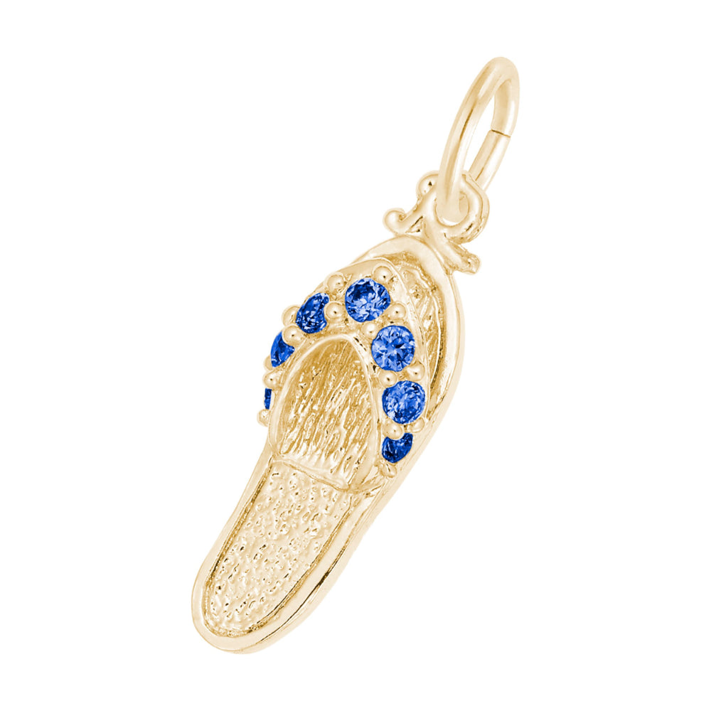 A gold charm shaped like a flip-flop sandal, featuring textured gold detailing and a row of round blue gemstones along the strap, set against a plain white background.