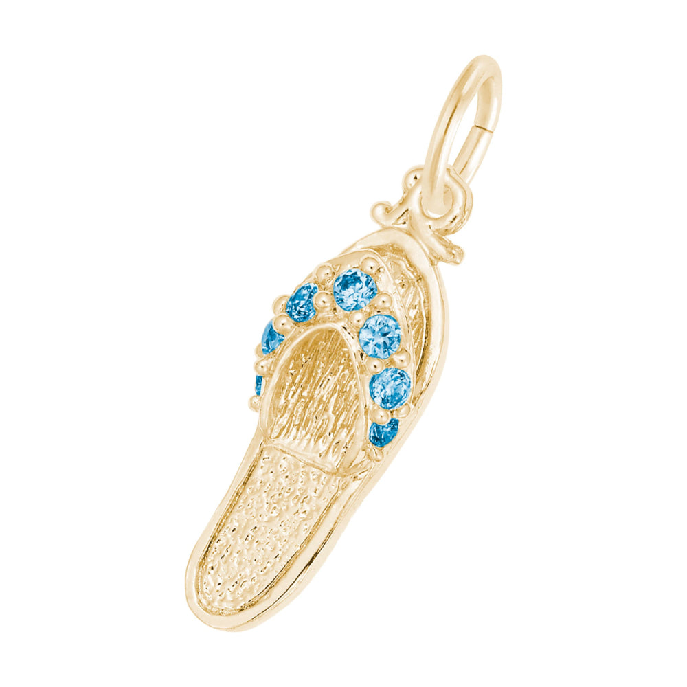 A gold charm shaped like a flip-flop sandal, featuring textured gold detailing and a row of round blue gemstones along the strap, set against a plain white background.