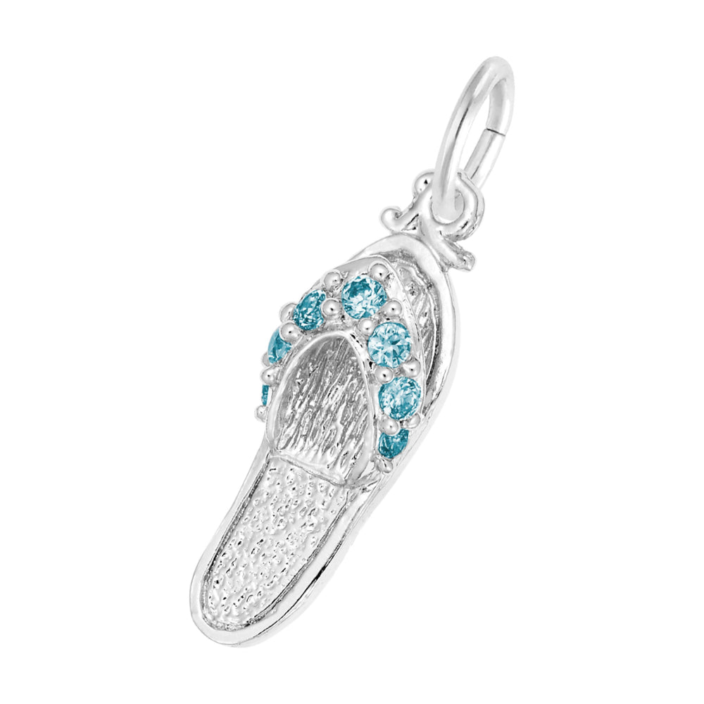 A silver charm shaped like a flip-flop sandal, featuring textured silver detailing and a row of round blue gemstones along the strap, set against a plain white background.