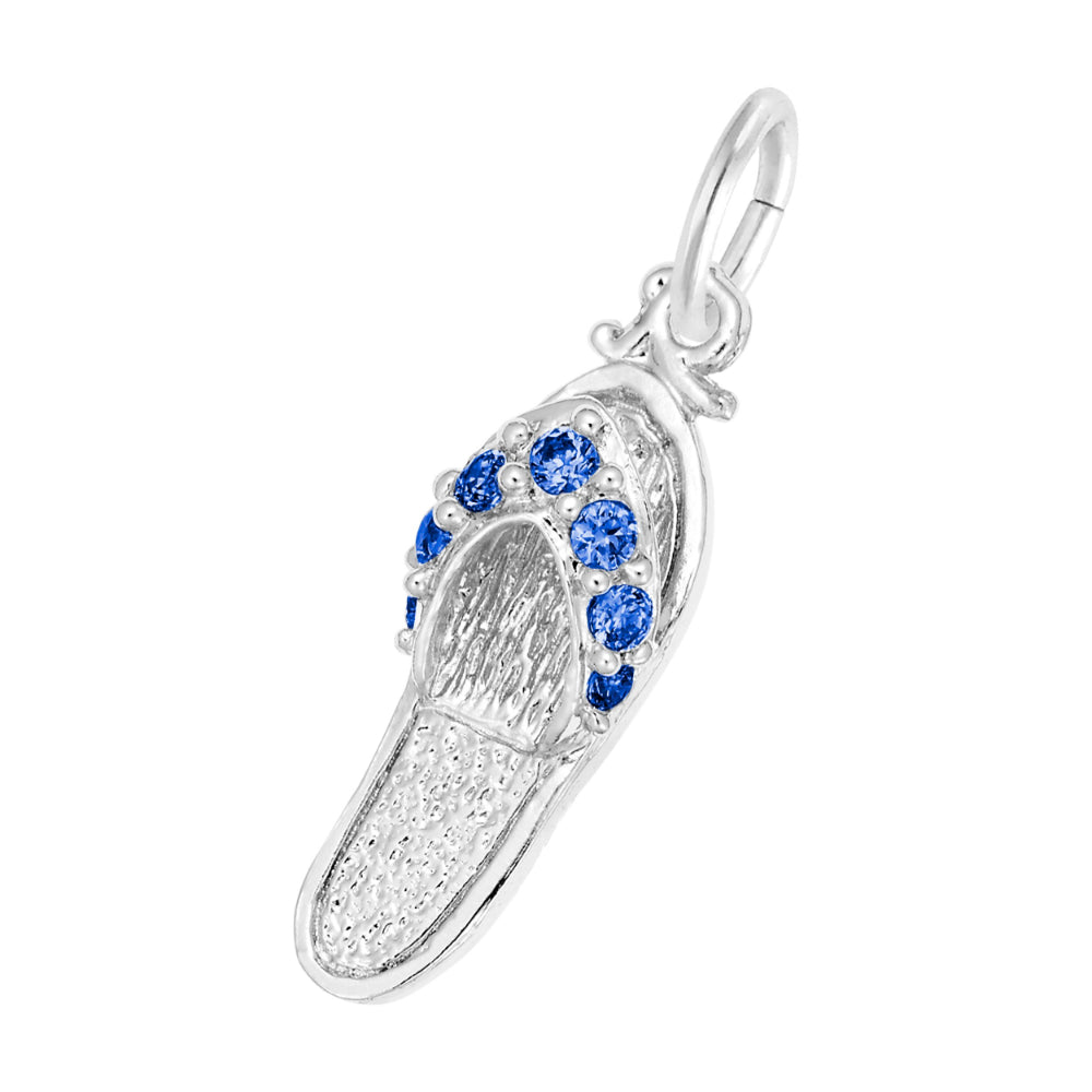 A silver charm shaped like a flip-flop sandal, featuring textured silver detailing and a row of round blue gemstones along the strap, set against a plain white background.