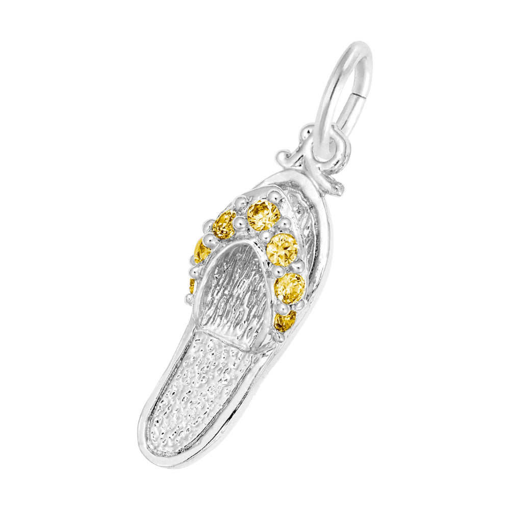 A silver charm shaped like a flip-flop sandal, featuring textured silver detailing and a row of round orange gemstones along the strap, set against a plain white background.