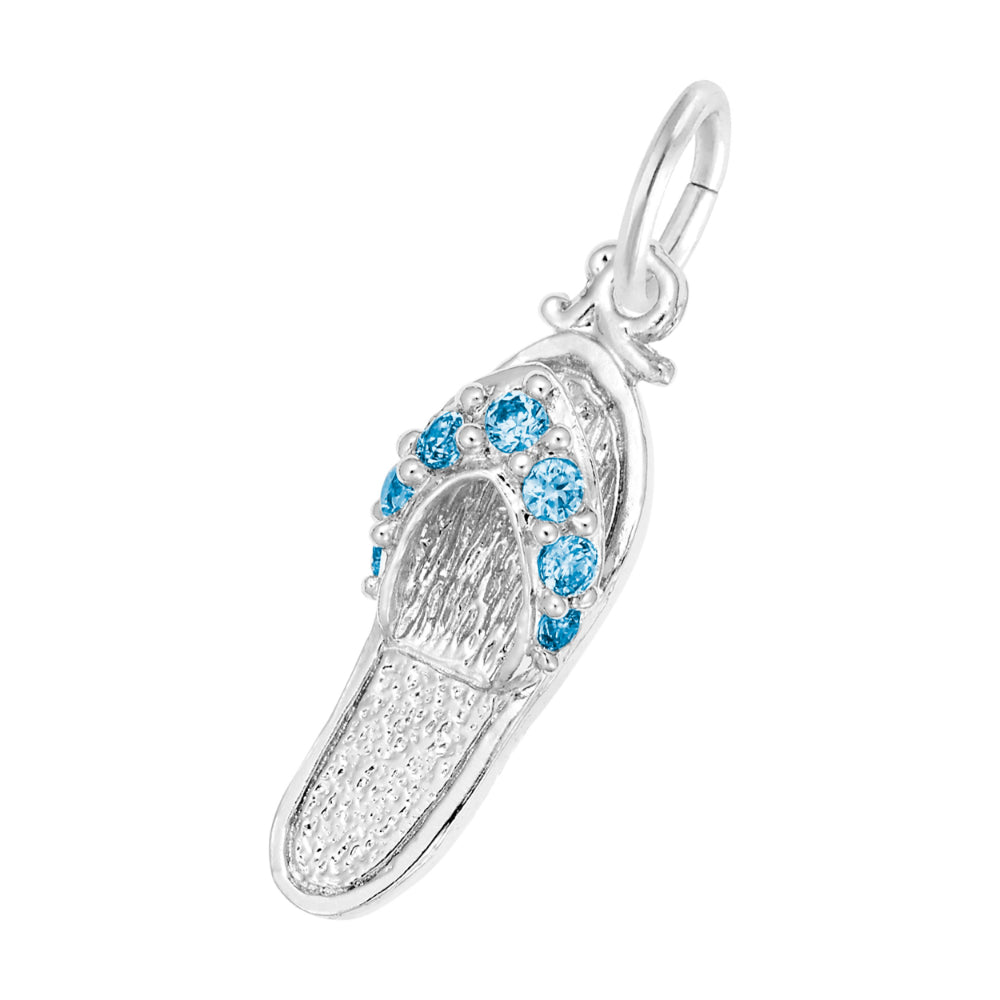 A silver charm shaped like a flip-flop sandal, featuring textured silver detailing and a row of round blue gemstones along the strap, set against a plain white background.