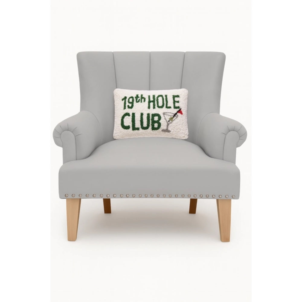 A light gray upholstered armchair with wooden legs and nailhead trim, featuring a white throw pillow that reads '19th HOLE CLUB' in green with a martini glass graphic.