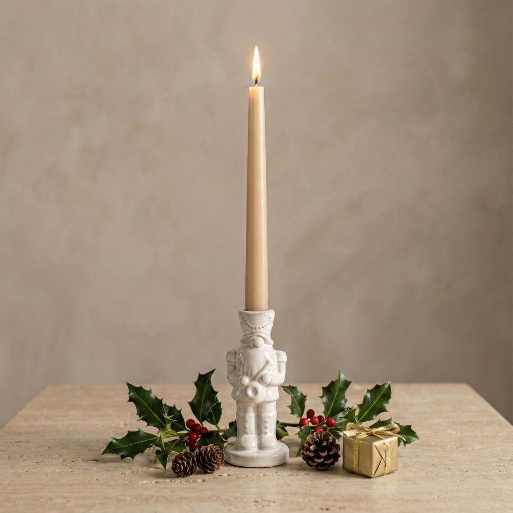 A lit cream taper candle in a white nutcracker-shaped holder, styled on a light stone surface with holly leaves, pinecones, and a small wrapped gift against a warm beige background.