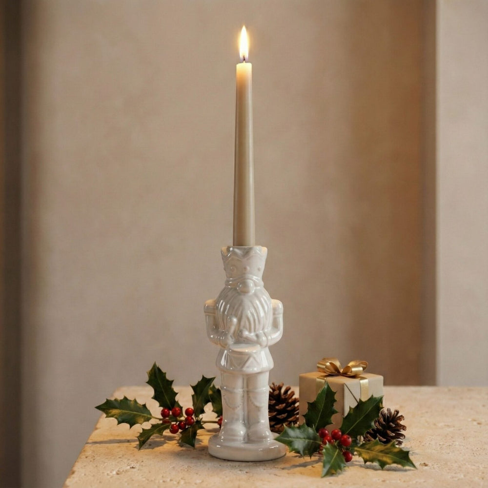 A white ceramic nutcracker candle holder with a lit taper candle, styled on a stone surface with holly, pinecones, and a small gift box against a neutral beige background.