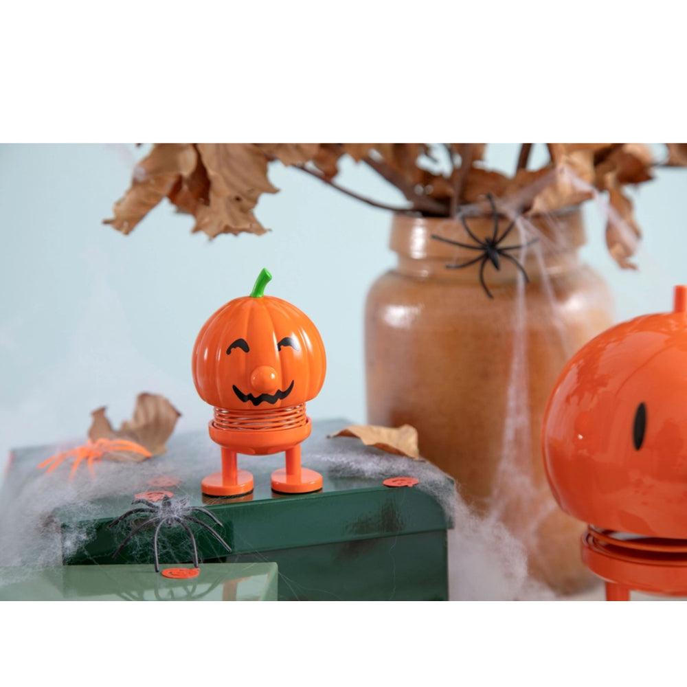An orange jack-o'-lantern-shaped spring toy with a carved pumpkin face and small green stem, displayed on a green box with Halloween props including fake spiderwebs, a brown jar, dried leaves, and a black plastic spider.