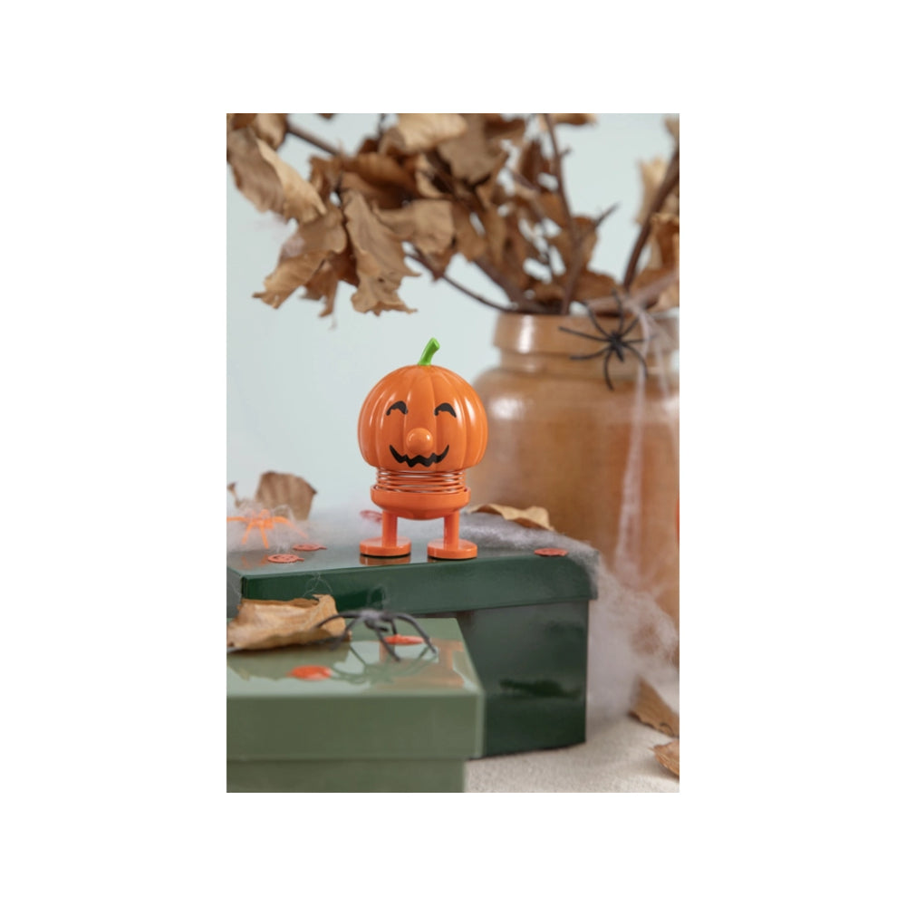 An orange pumpkin-shaped toy with a smiling jack-o'-lantern face and small legs stands on a green box in a styled Halloween scene with dried leaves, a jar, and spider decorations.