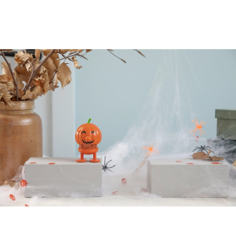 An orange jack-o'-lantern-shaped figurine with a smiling face stands on a white display block in a Halloween-themed setup with fake spiderwebs, a brown vase with dried leaves, and plastic spiders.