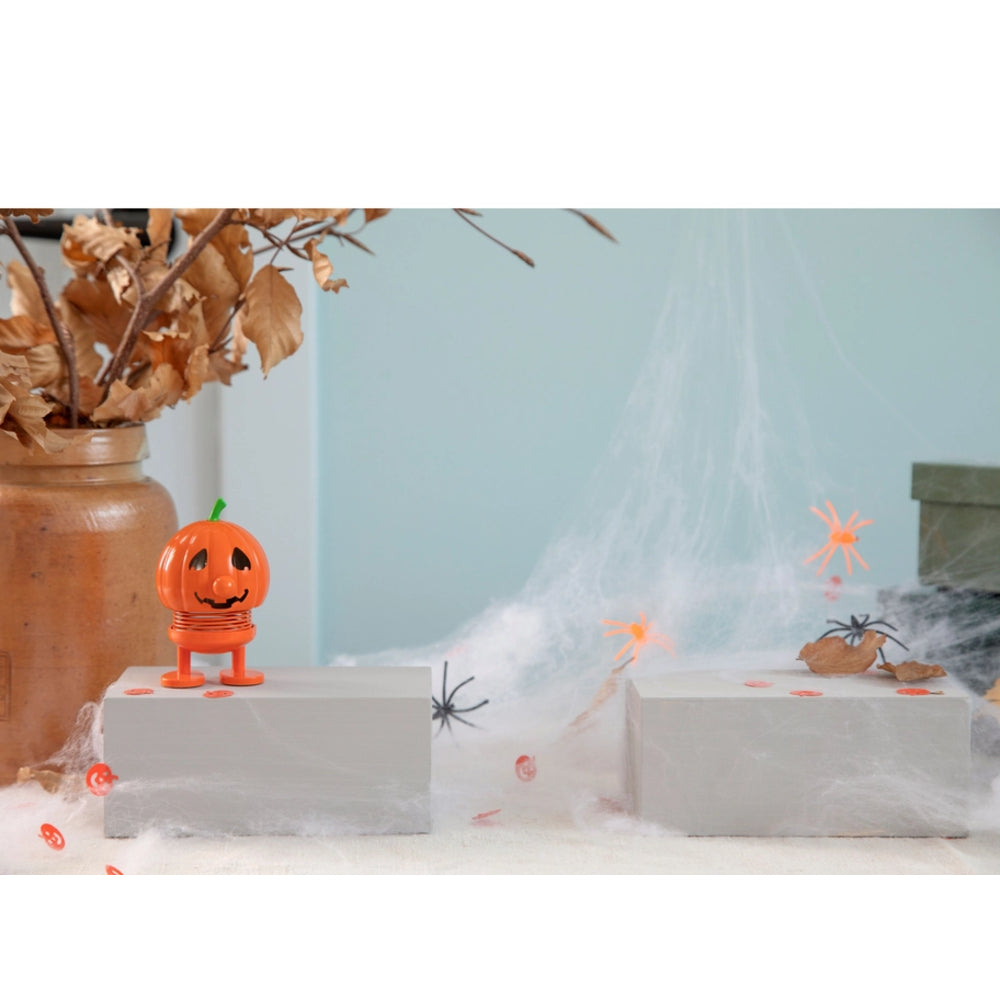 An orange jack-o'-lantern toy figure with a green stem stands on a white display block in a Halloween-themed setup with fake cobwebs, plastic spiders, dried leaves in a jar, and a pale blue background.