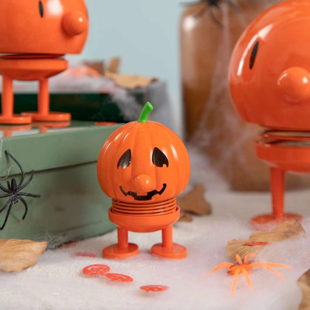 Small orange jack-o'-lantern toy with legs on a festive background with pumpkins and spider decorations.