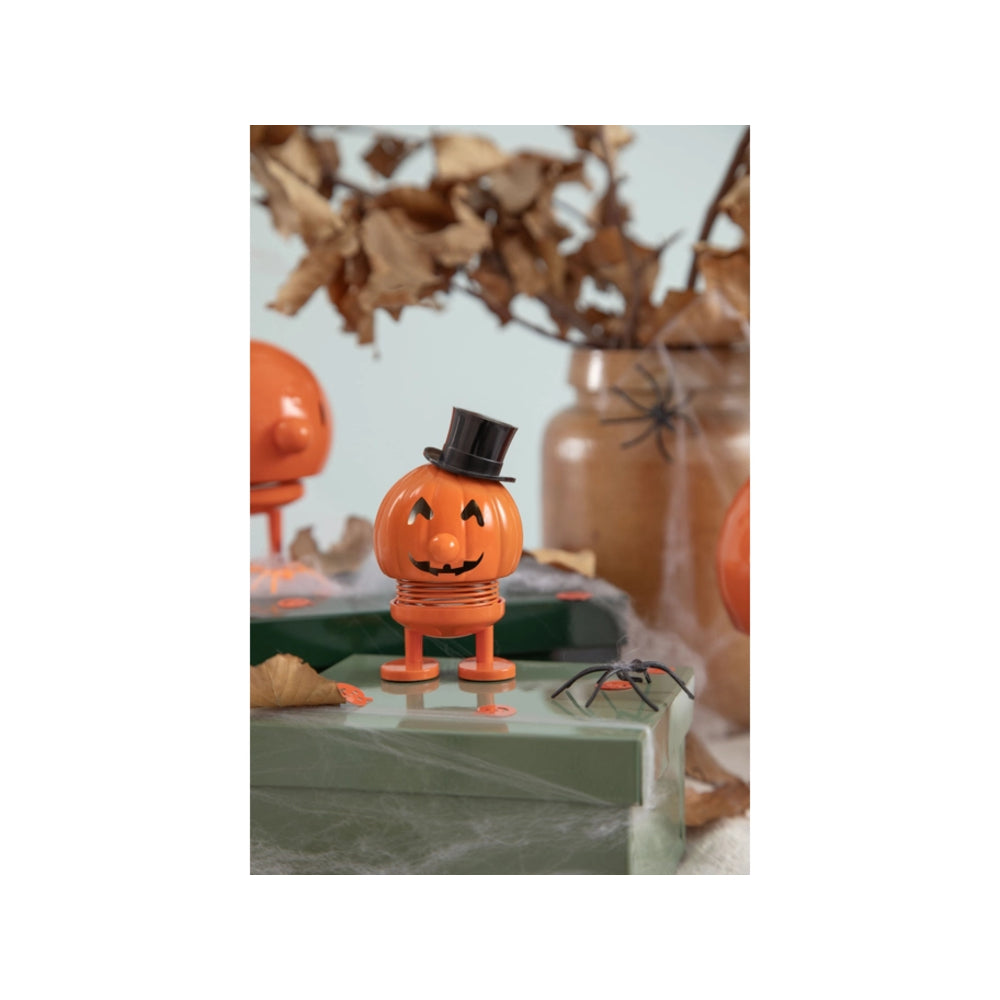 An orange jack-o'-lantern Halloween decoration with a carved face, small legs, and a black top hat, standing on a tabletop with spider and dried leaves in the background.