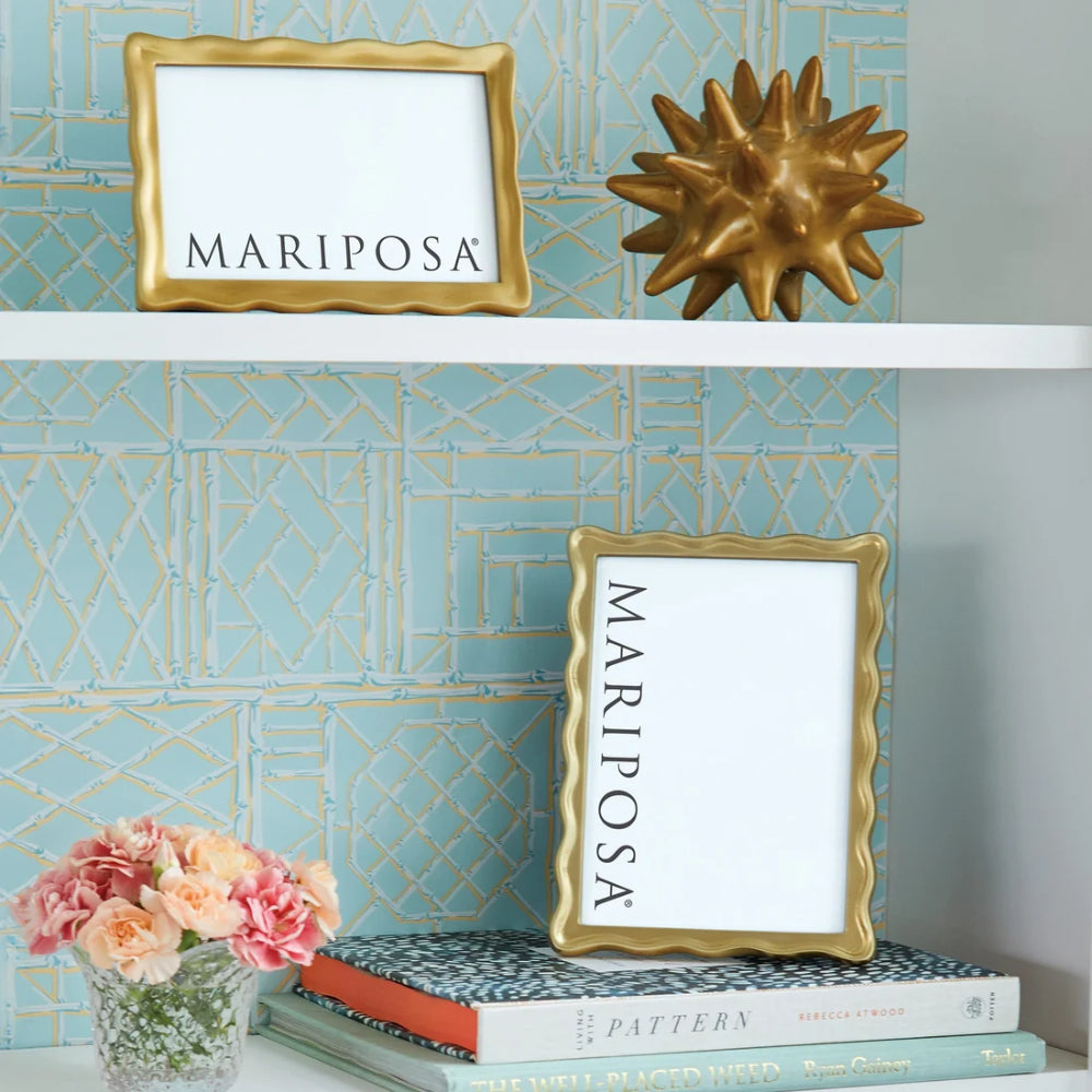 Decorative shelf with gold 'Mariposa' frames, books, and flowers against a patterned wall.