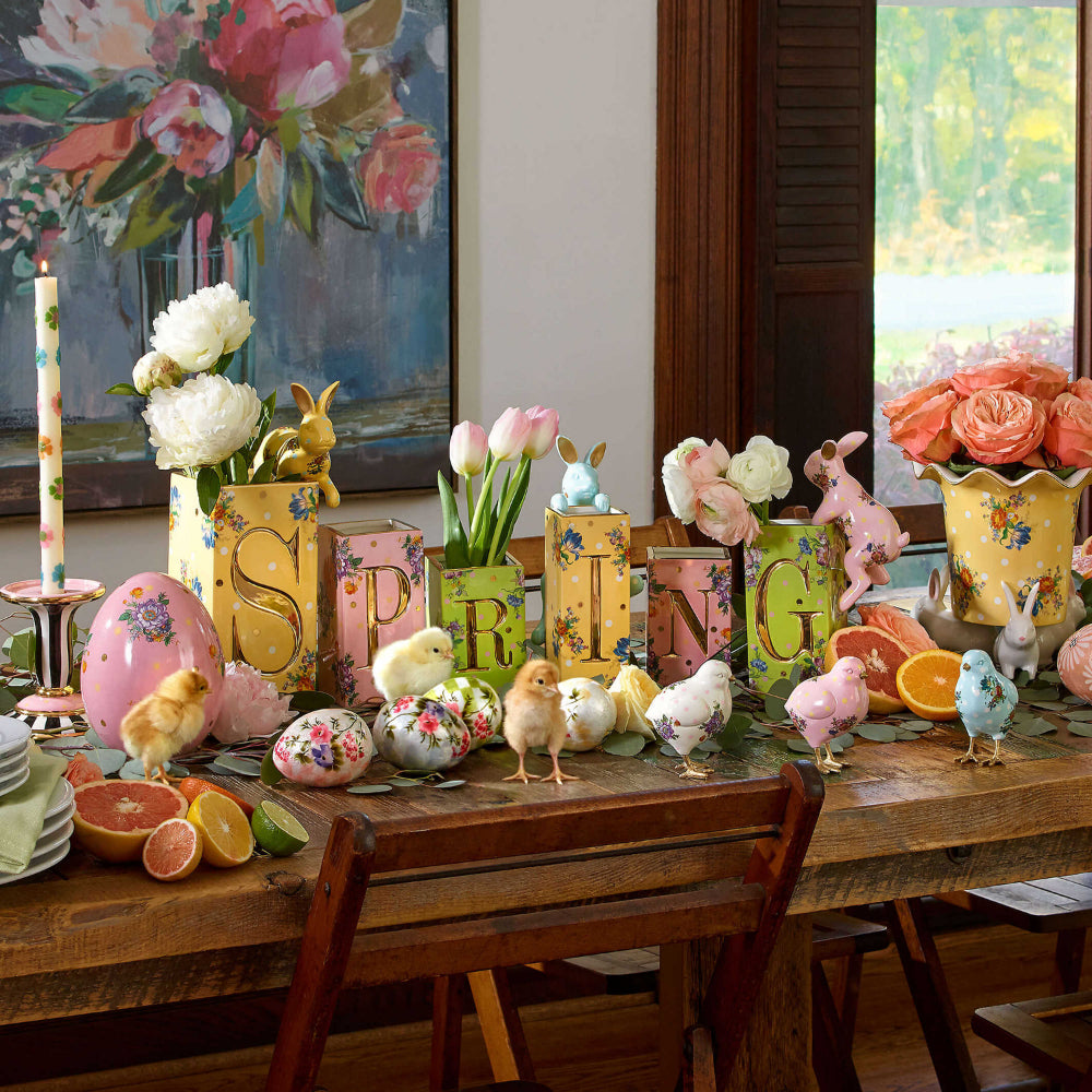 A spring multi-colored life style photo of MacKenzie-Childs mod flower dinner candles on a table with flowers and bunnies and  that says "SPRING"