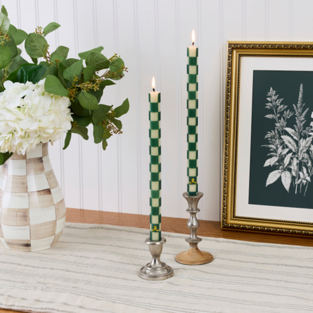 Two lit taper candles with a green and cream checkered pattern, displayed in silver and wooden candlesticks on a table runner, with a vase of white flowers and a framed botanical print in the background.