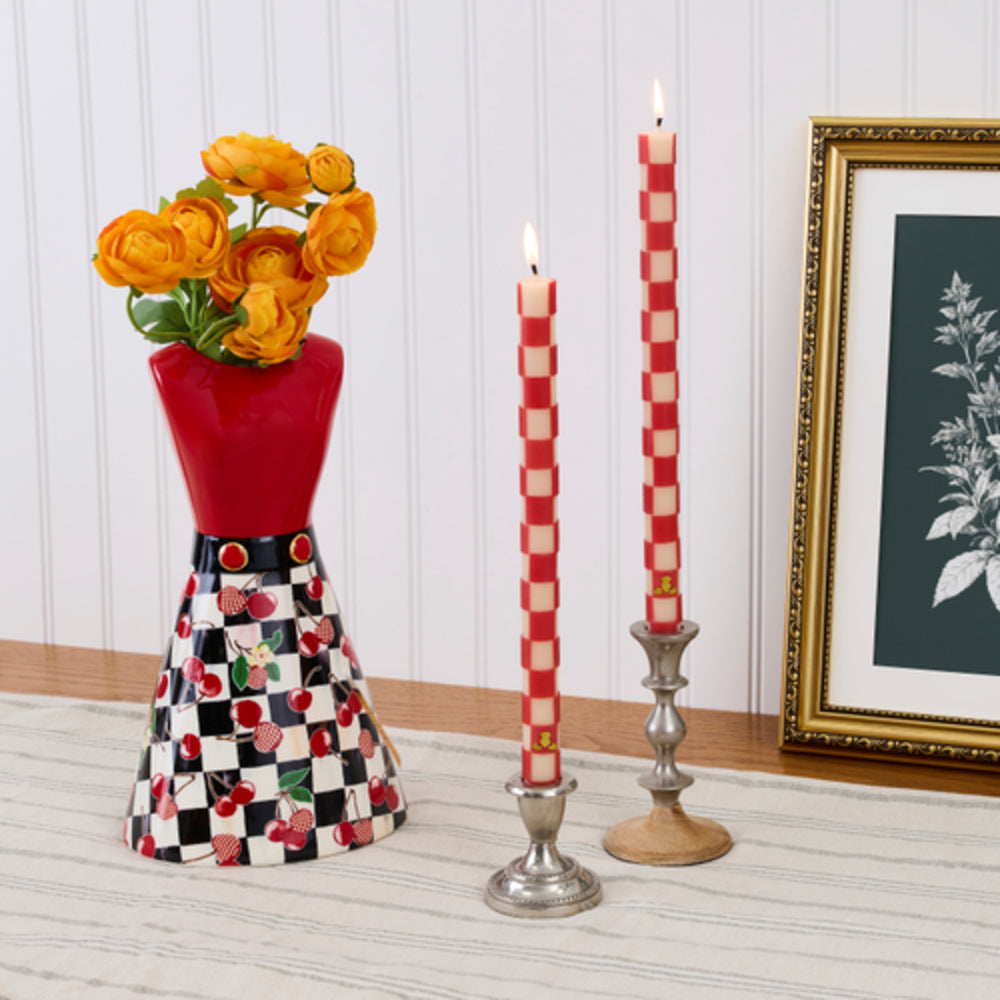 A decorative ceramic vase with a red upper section and a black-and-white checkerboard lower section decorated with cherries, holding orange flowers. Two red-and-white checkered taper candles in silver and wood candlesticks are placed beside it on a light wood surface against a white paneled wall, with a framed botanical print partially visible on the right.