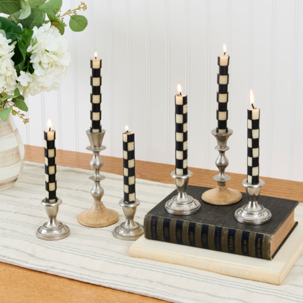 Six black and cream checkered taper candles in silver candlesticks, styled on a table runner with vintage books and a floral arrangement.