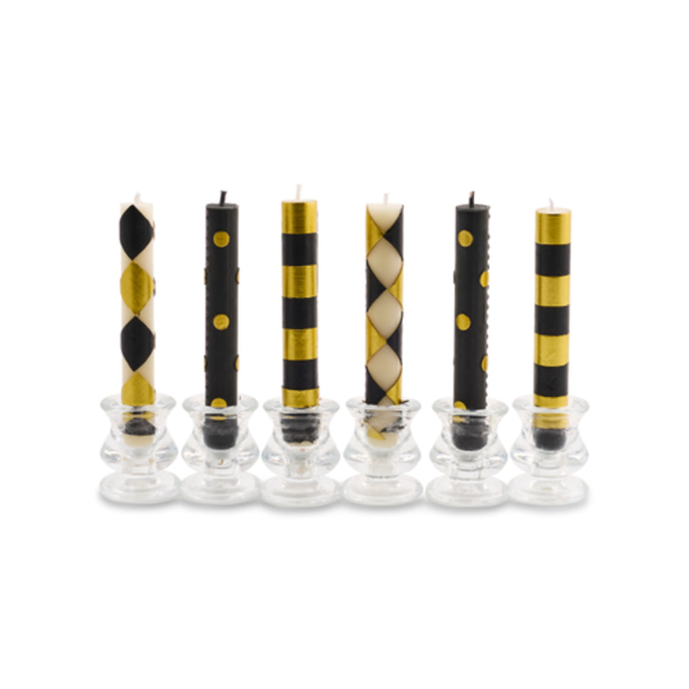 Six patterned taper candles in clear glass holders on a white background. The candles feature black and gold designs, including stripes, harlequin diamonds, and polka dots.