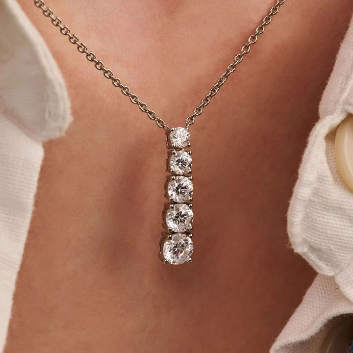 A close-up of a silver-toned necklace with a vertical drop pendant featuring five clear round-cut stones, worn on a person's neck over a white collared shirt.