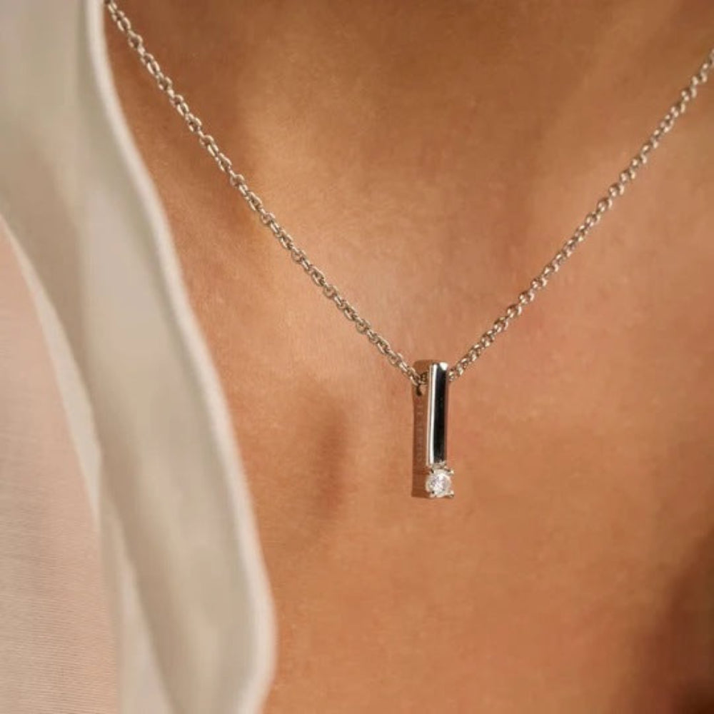 A close-up of a delicate silver necklace with a vertical bar pendant and a small clear gemstone, worn on a model's neck.