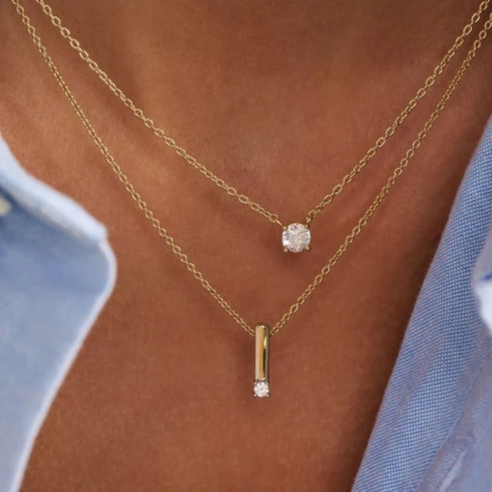 A close-up of a person's neck and chest wearing two layered gold necklaces. The shorter necklace has a round clear gemstone pendant, and the longer necklace has a slim rectangular gold bar pendant with a small clear gemstone at the bottom.