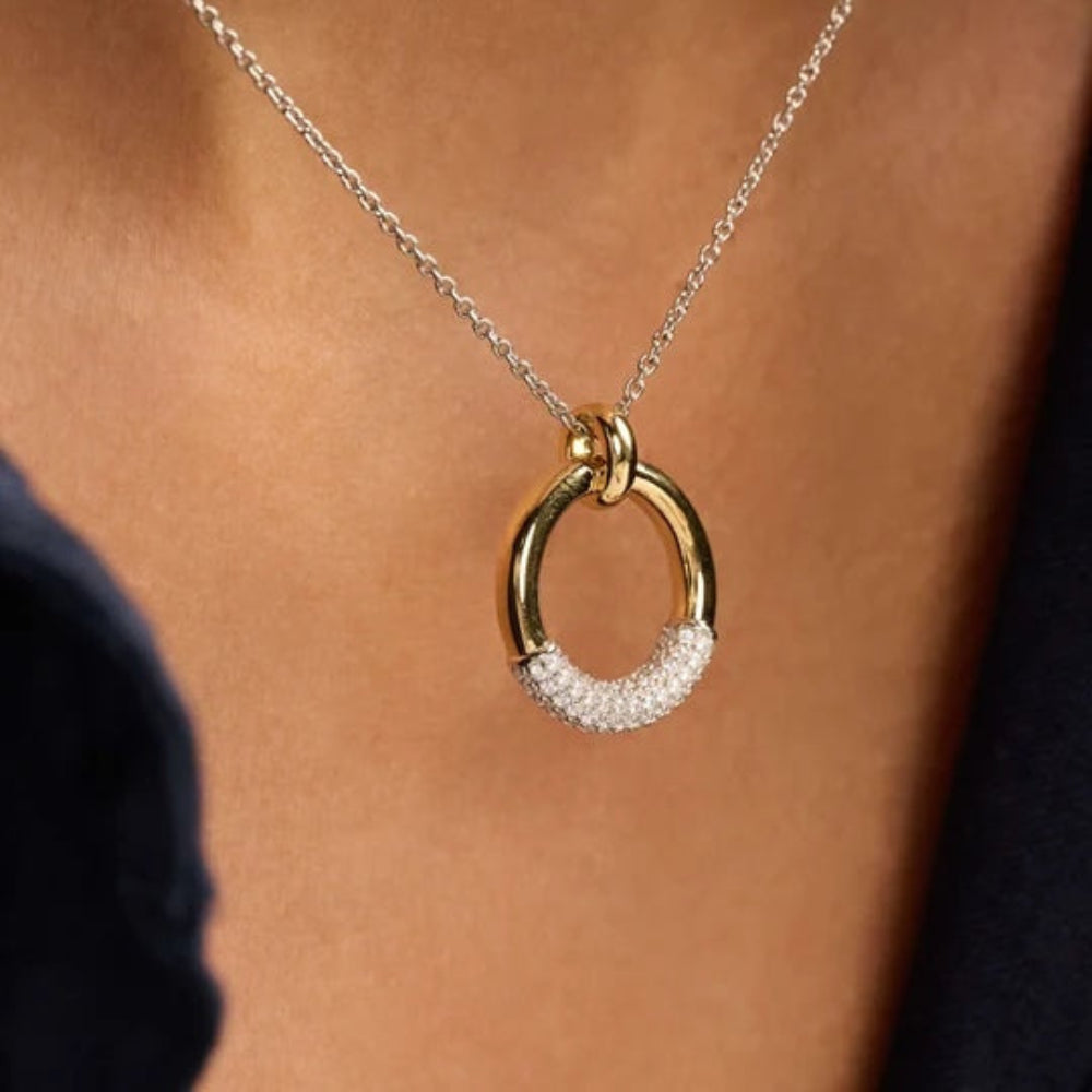 A close-up of a gold and silver necklace with a half-circle pendant, worn on a model's neck. The lower half of the pendant is set with small clear pavé stones.