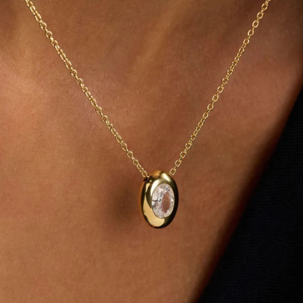 A gold chain necklace with an oval pendant featuring a clear faceted gemstone, shown worn on a person's neck against a dark background.