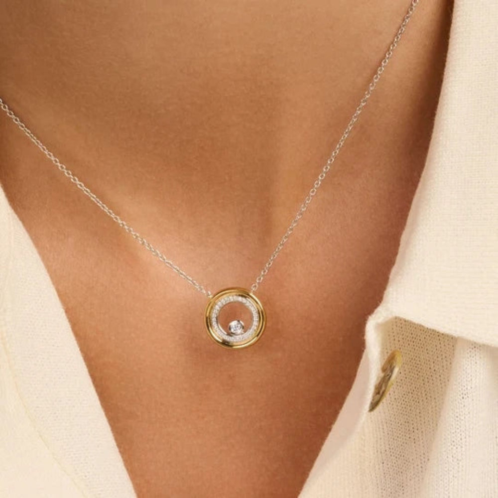 A close-up of a person wearing a delicate necklace with a gold and silver double-ring pendant set with a small clear gemstone, styled with a cream-colored collared shirt.