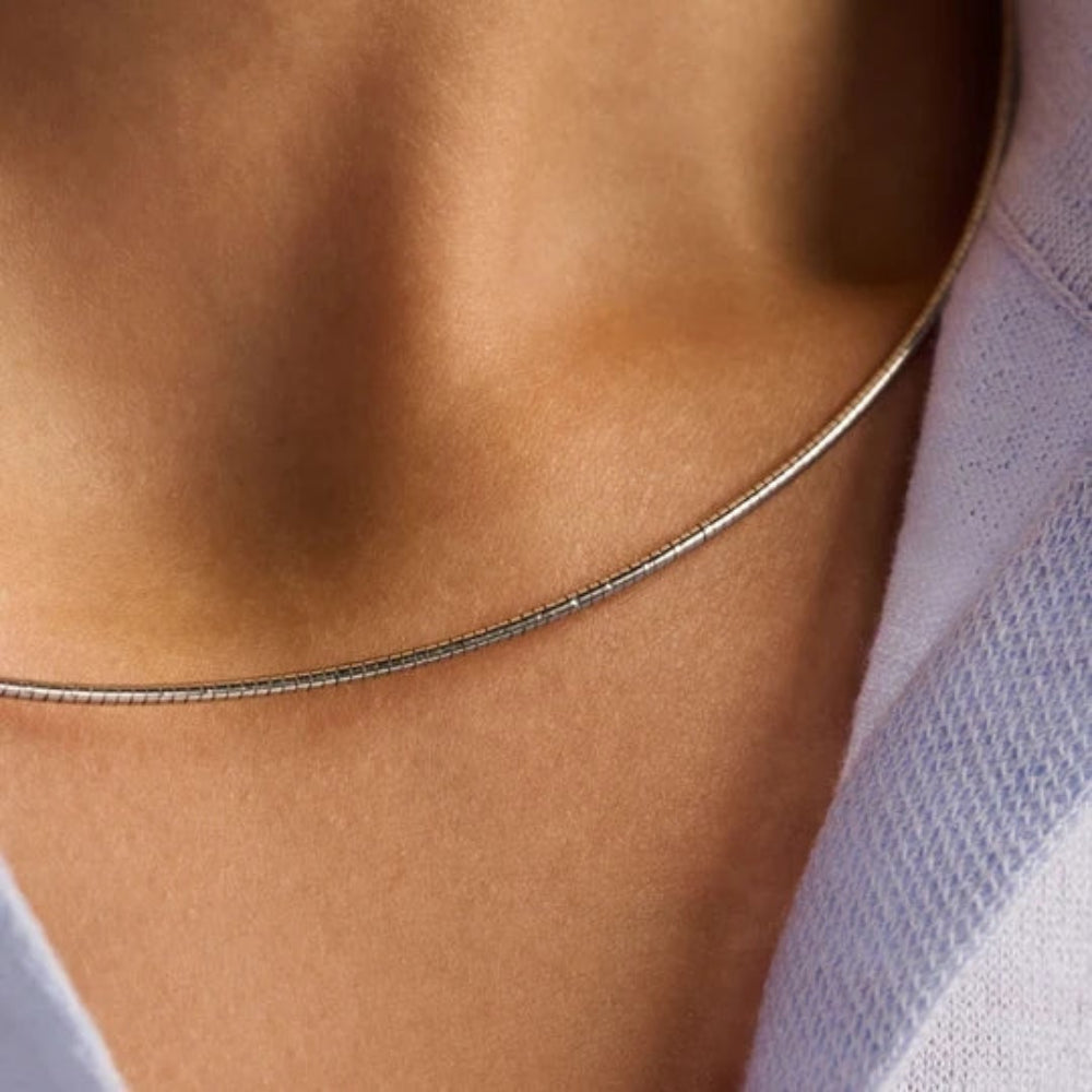 A close-up of a delicate gold snake chain necklace worn on a person's neck, paired with a light lavender knit top.