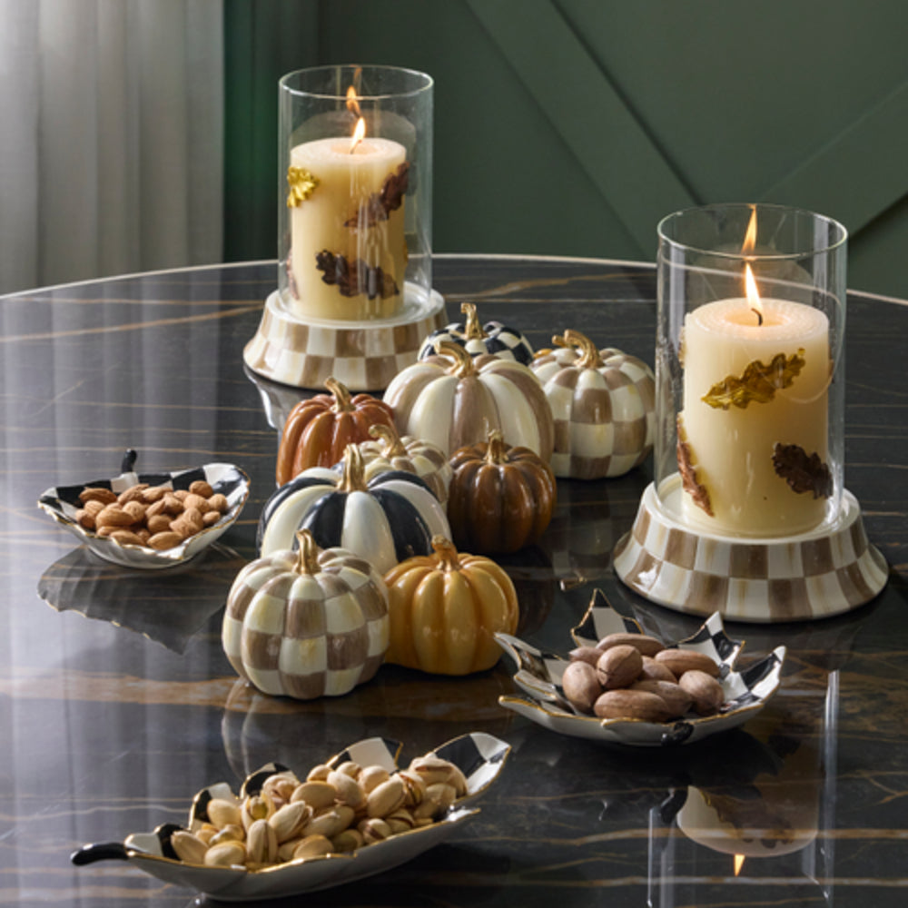 Two lit pillar candles in clear glass cylinders with cream wax and dried botanical inclusions, displayed on a checkered base on a dark marble table. Decorative pumpkins and bowls of nuts are arranged around them in a styled indoor setting.