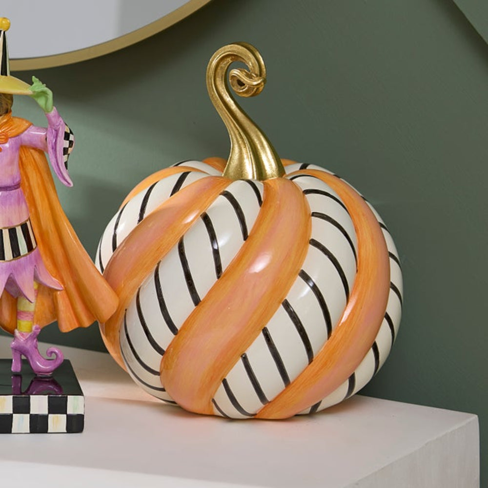 A decorative pumpkin sculpture with a glossy white body, black vertical stripes, and wide peach-orange bands, topped with a metallic gold stem. It is displayed on a white surface against a muted green wall, next to a small witch figurine.