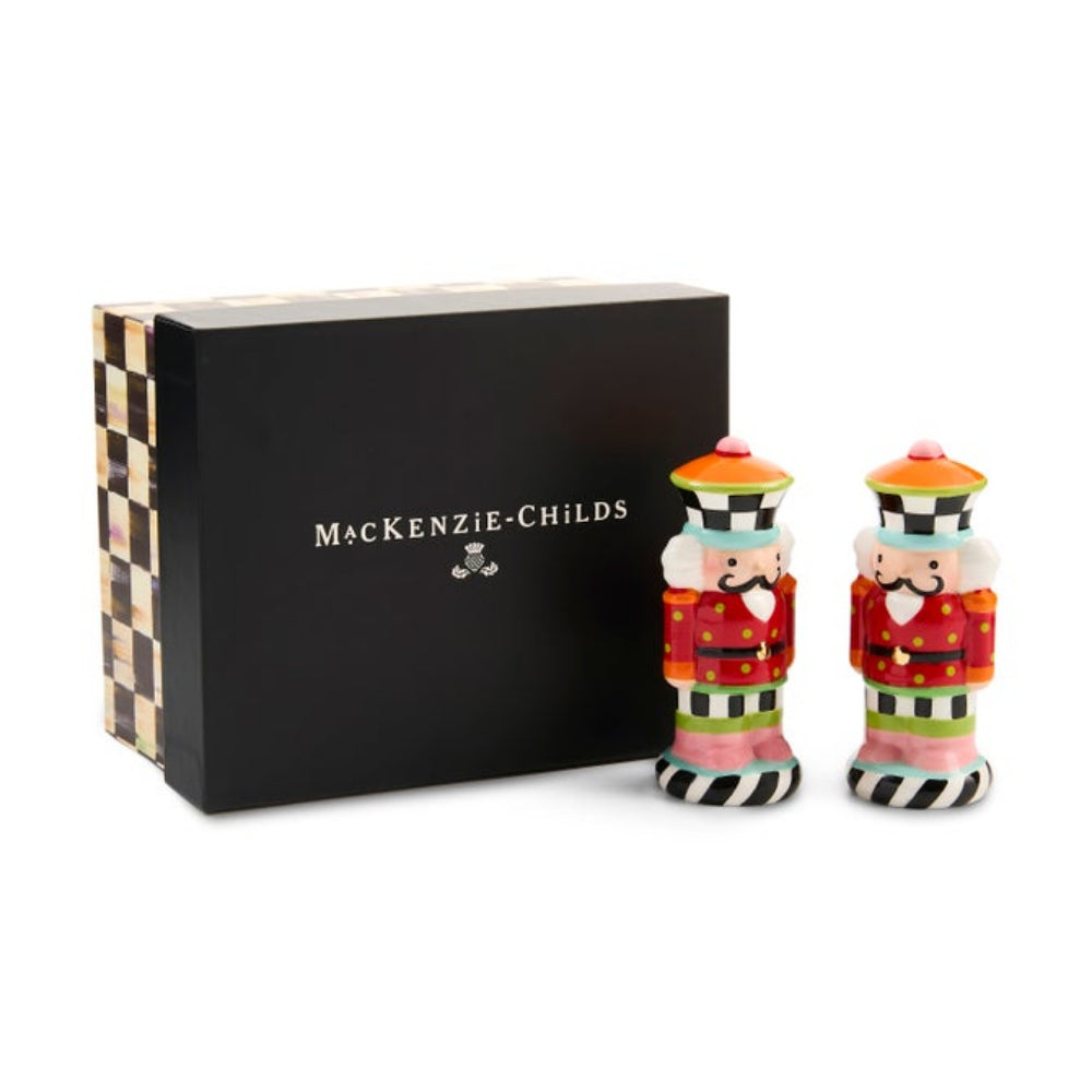 A pair of MacKenzie-Childs nutcracker salt and pepper shakers standing in front of their black presentation box. The shakers are decorated with vibrant, multicolored patterns including red, green, white, black, and gold, while the box features a black and gold checkered pattern.