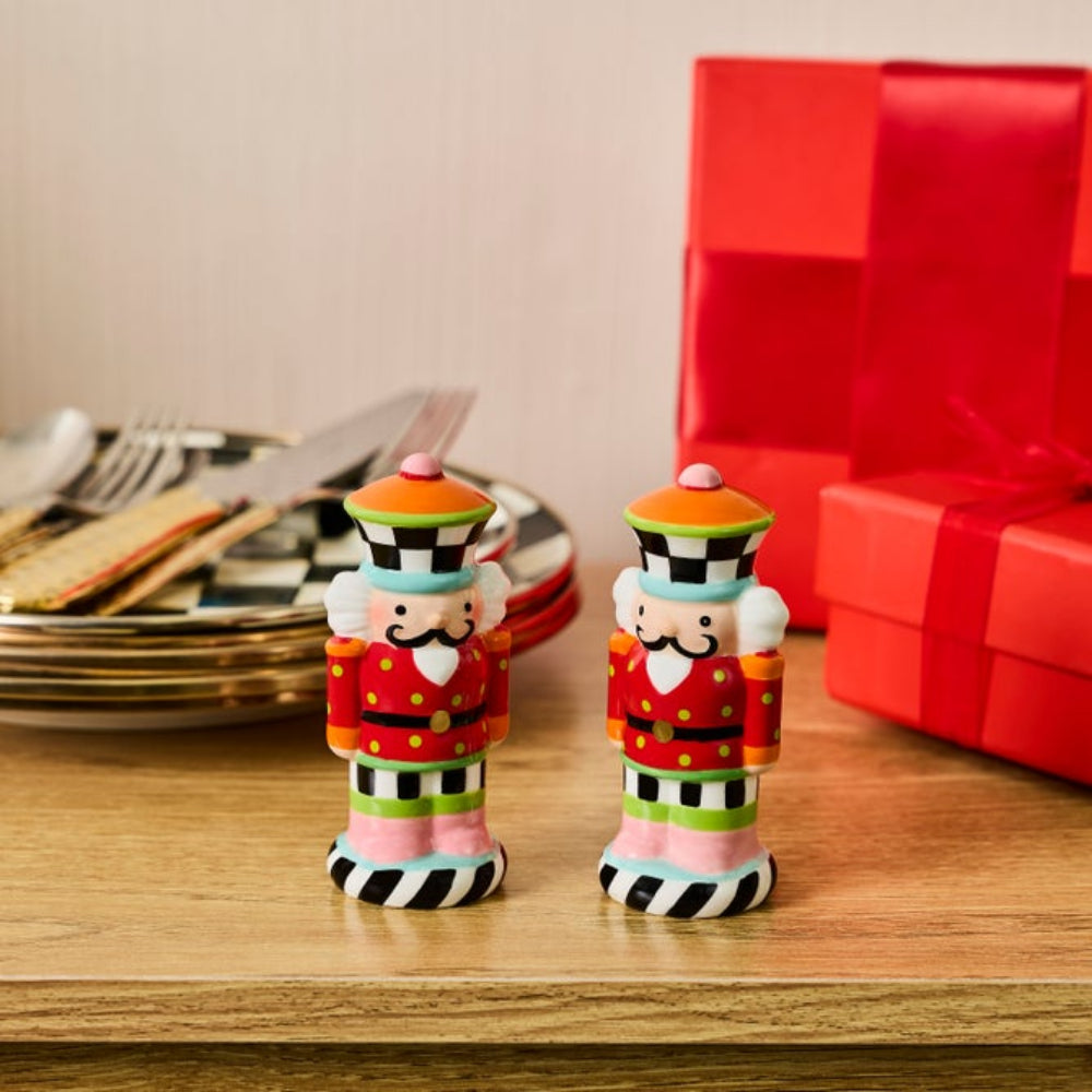 Two colorful nutcracker-shaped salt and pepper shakers standing on a wooden table, with plates and cutlery on the left and red gift boxes on the right.