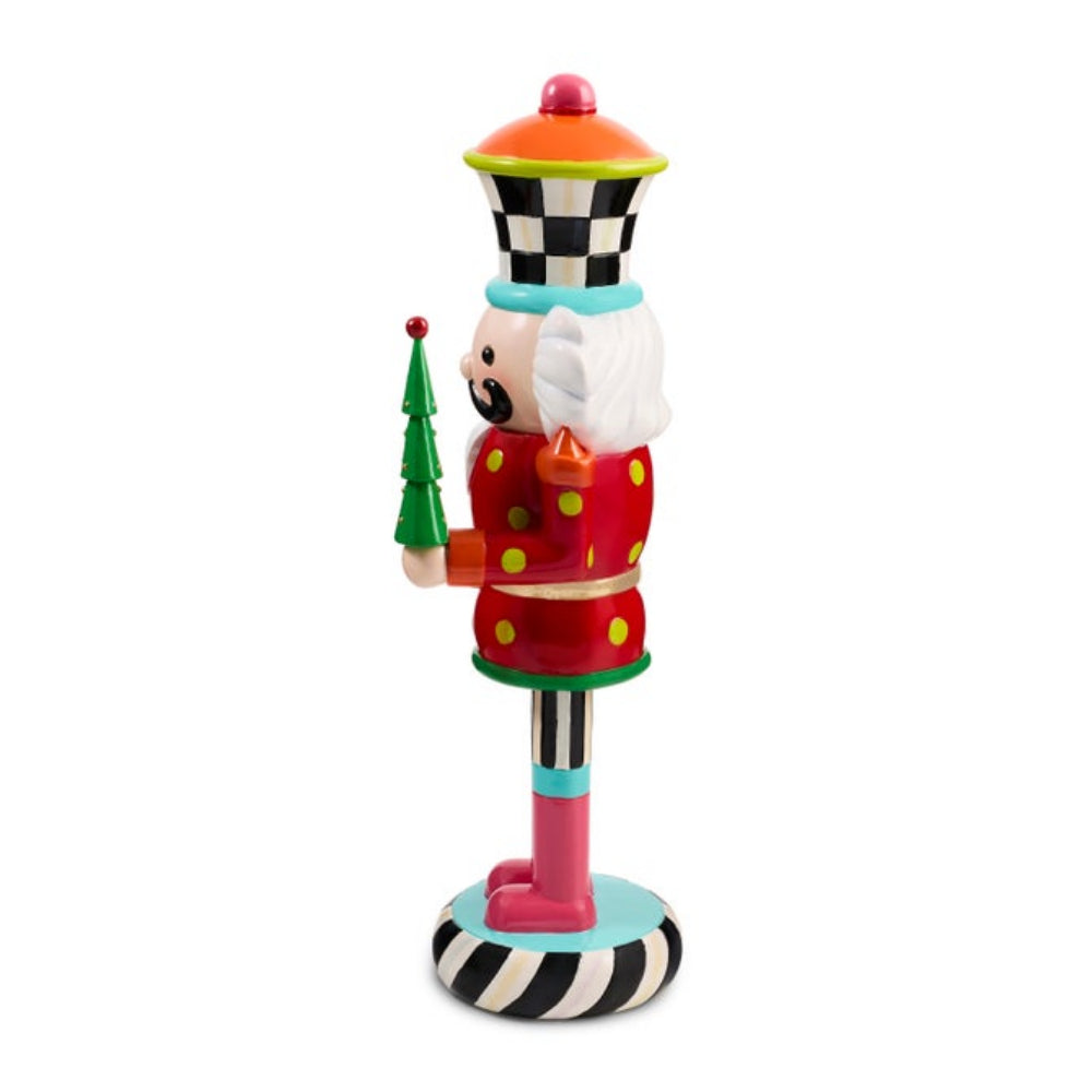 A whimsical, colorful nutcracker figurine holding a small green Christmas tree. The nutcracker features a red polka-dot coat, a black-and-white checkered hat with a red top, a white beard, and a pink base with black-and-white stripes, set against a plain white background.
