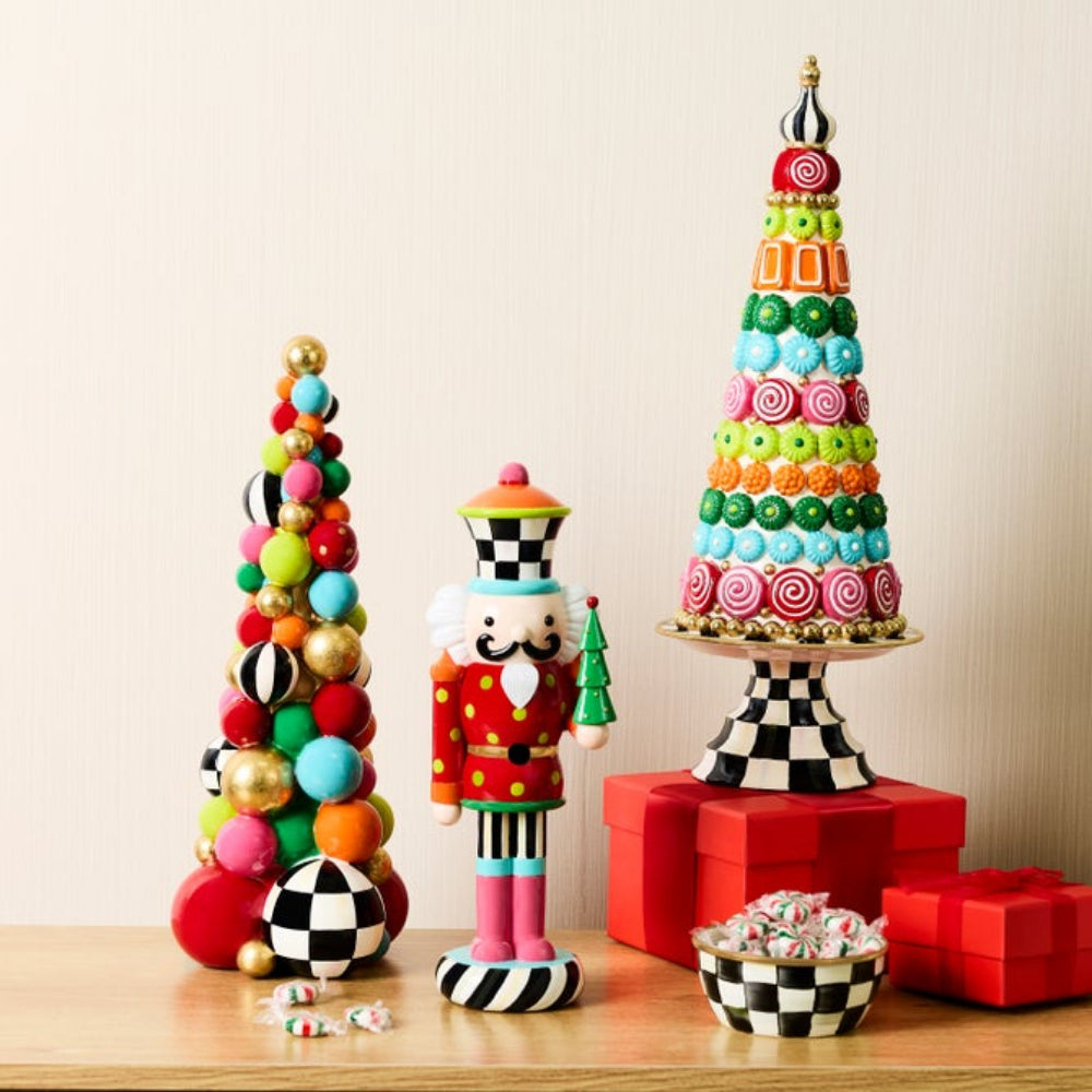 A festive tabletop display featuring a colorful candy tree on a pedestal, a nutcracker figurine, and red gift boxes on a light wood surface against a cream wall.